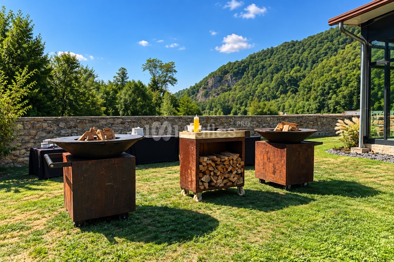 Espace extérieur avec foyers en métal, bois de chauffage et brasero, montagnes boisées en arrière-plan.
