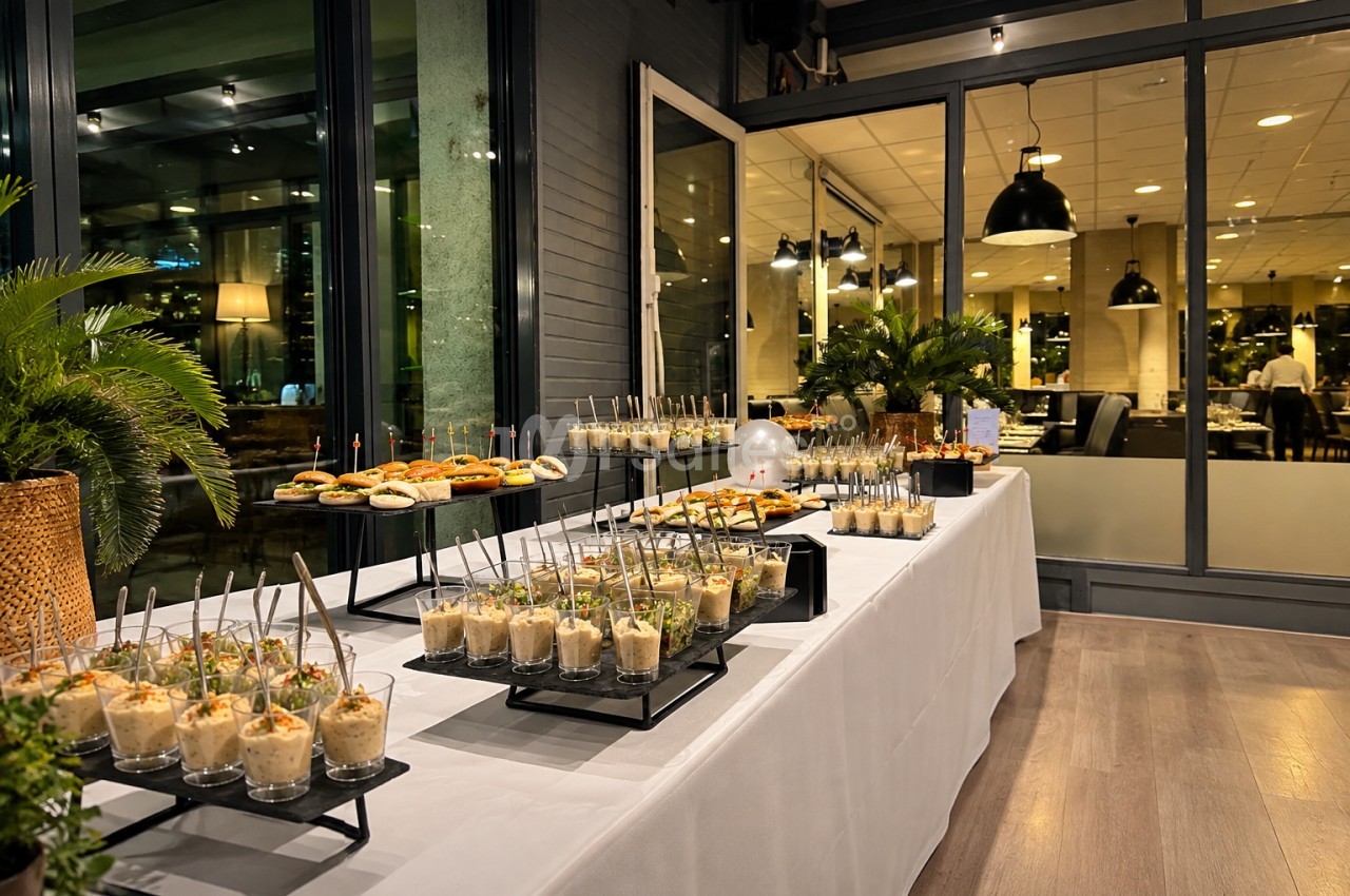 Buffet dressé avec mises en bouche et verres, tables blanches, baies vitrées ouvertes sur salle intérieure éclairée.