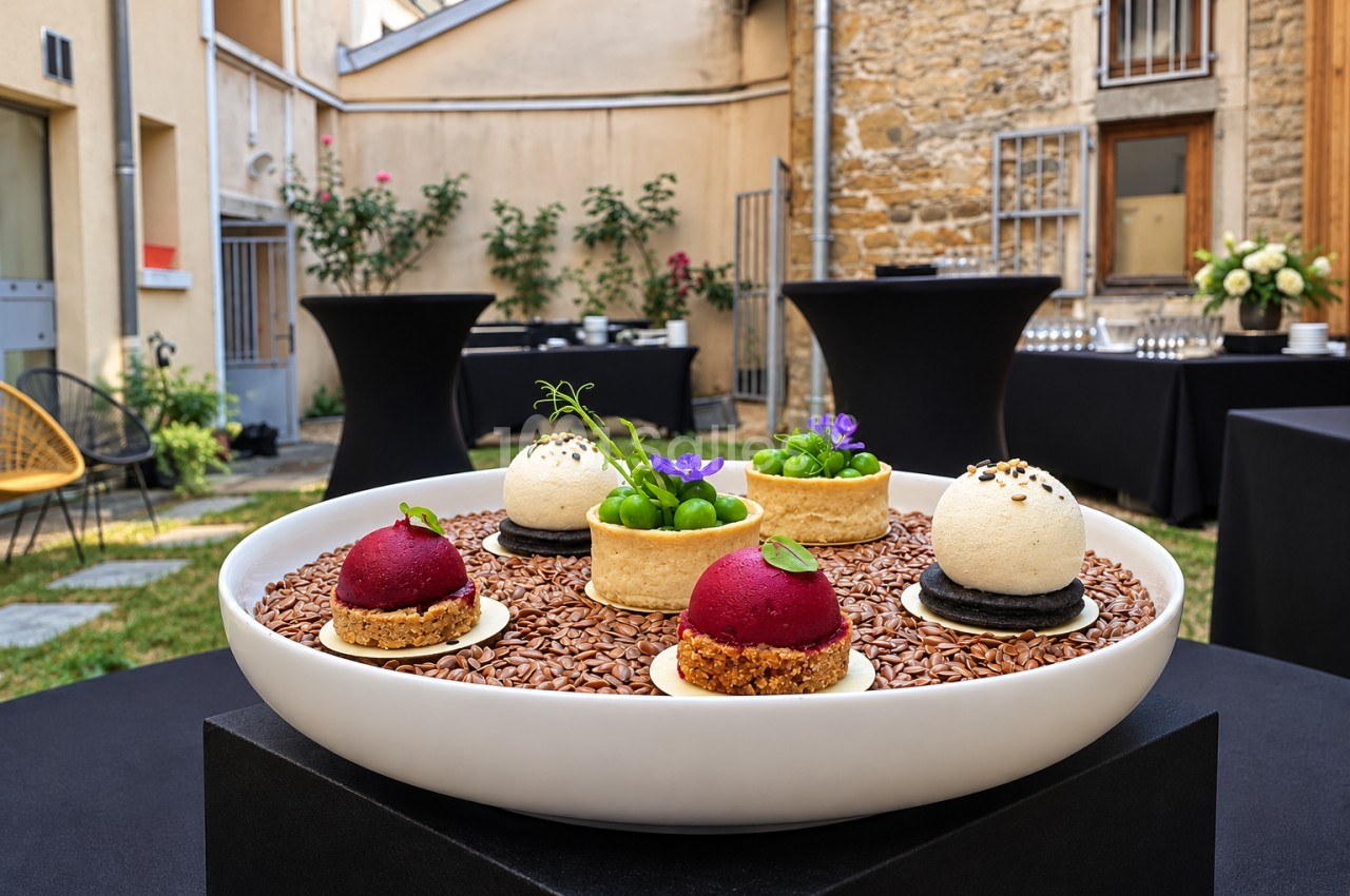 Présentation de desserts gastronomiques sur lit de graines dans une cour pavée avec tables hautes noires et façades en pierre.