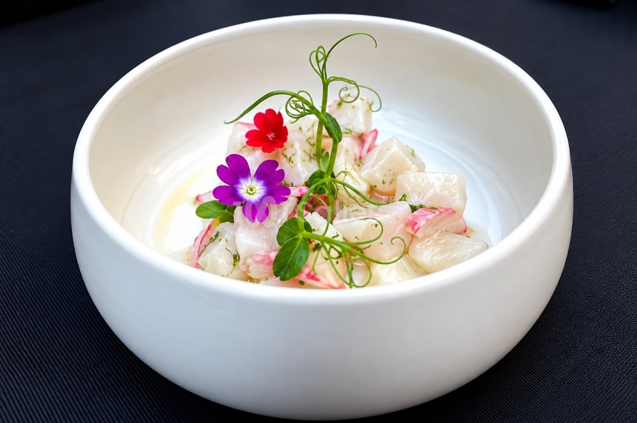 Bol blanc contenant des dés de poisson blanc, fleurs comestibles rouges et violettes, micropousses vertes et filet de sauce.