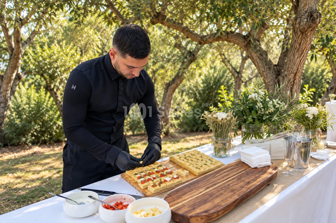 Traiteur en noir dressant des canapés sur une table extérieure sous les arbres, entouré de verres et de fleurs.