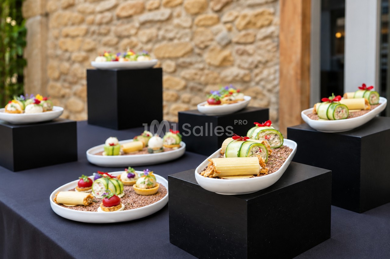 Assortiment de bouchées dressées sur des socles noirs - roulés de concombre, canapés et petits fours devant un mur en pierre.