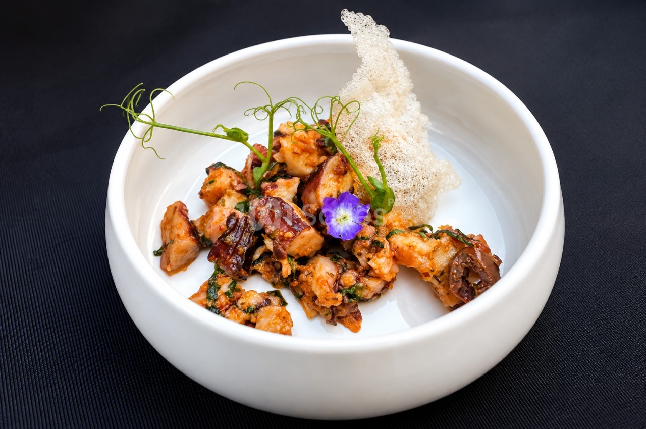 Assiette blanche avec morceaux de viande caramélisée, tuile croustillante, germes et fleur violette.