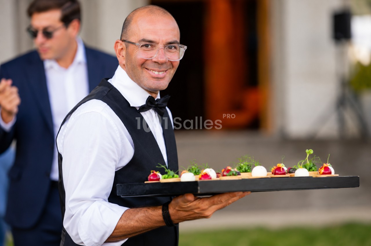 Serveur en costume présentant un plateau de petits fours sur canapés aux fruits rouges et herbes fraîches.