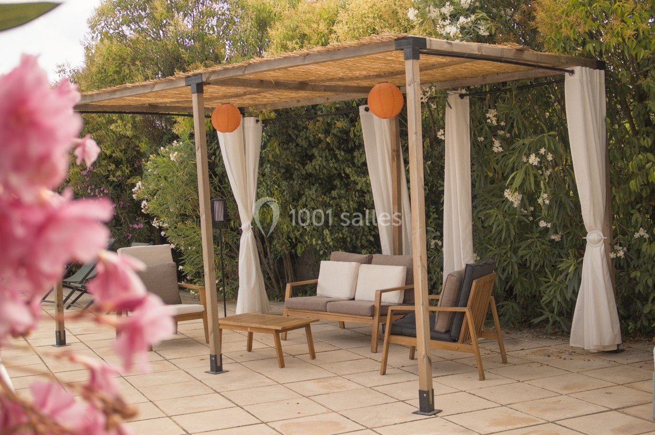 Pergola en bois avec rideaux blancs, mobilier de jardin et lanternes suspendues, entourée de végétation fleurie.