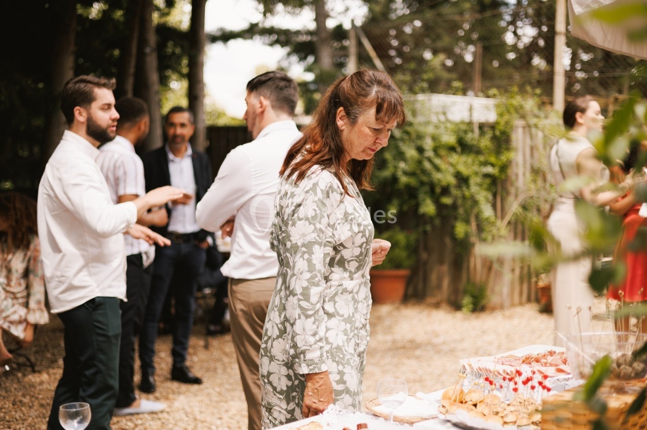 Une femme regarde un buffet en extérieur, tandis que des personnes discutent en arrière-plan dans un jardin.