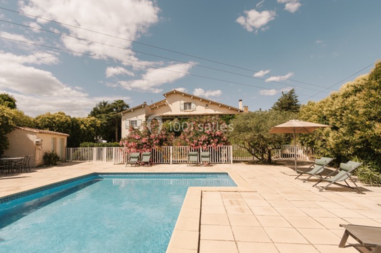 Maison avec jardin, piscine, transats et parasol, entourée de végétation sous un ciel partiellement nuageux.