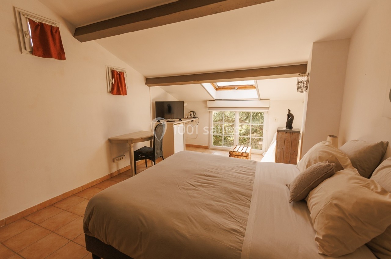 Chambre mansardée lumineuse avec lit double, bureau, télévision et fenêtre donnant sur un espace verdoyant.