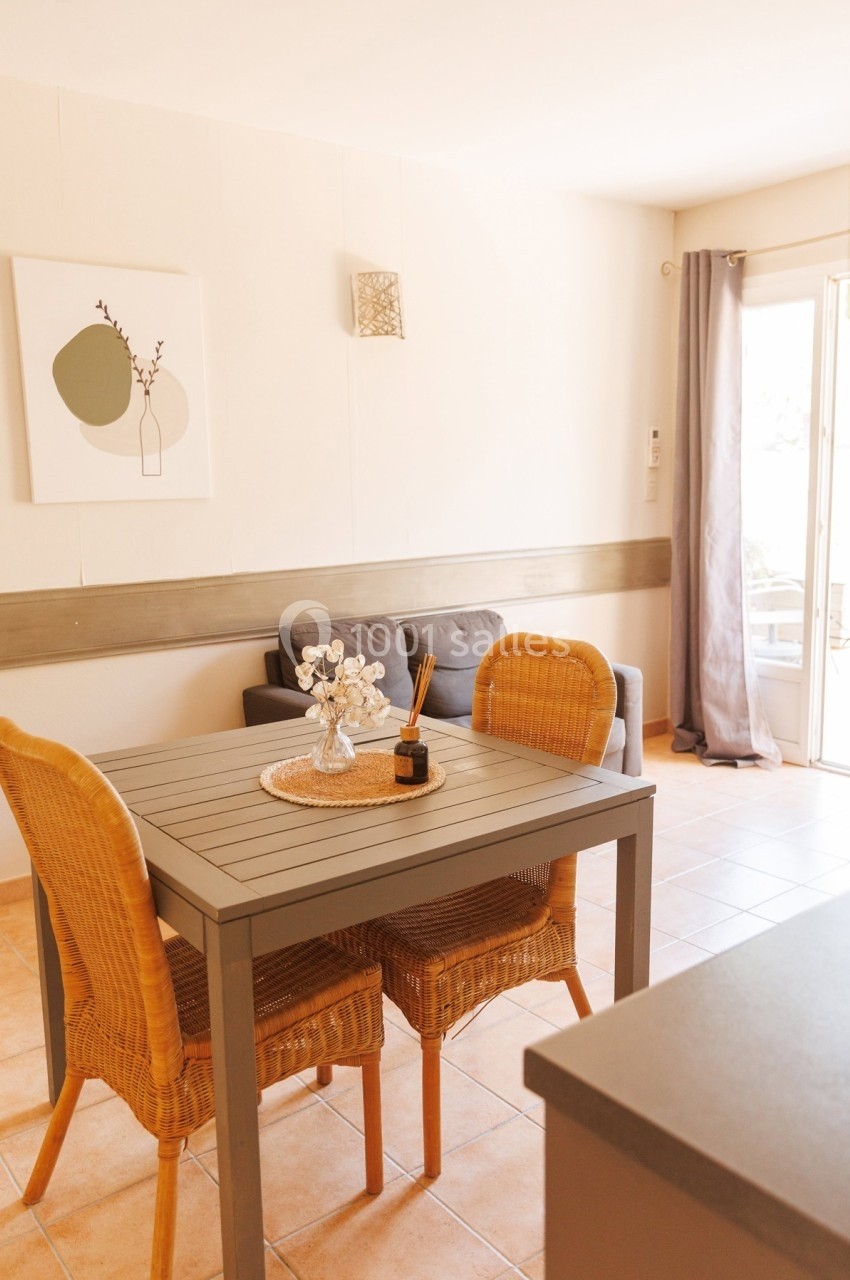 Salle à manger lumineuse avec table en bois, chaises en rotin, canapé gris et accès à une terrasse par une porte vitrée.