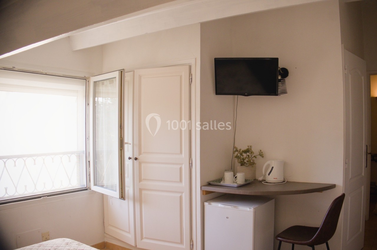 Chambre lumineuse avec fenêtre ouverte, petit réfrigérateur, bouilloire, télévision murale et chaise près d'une table.