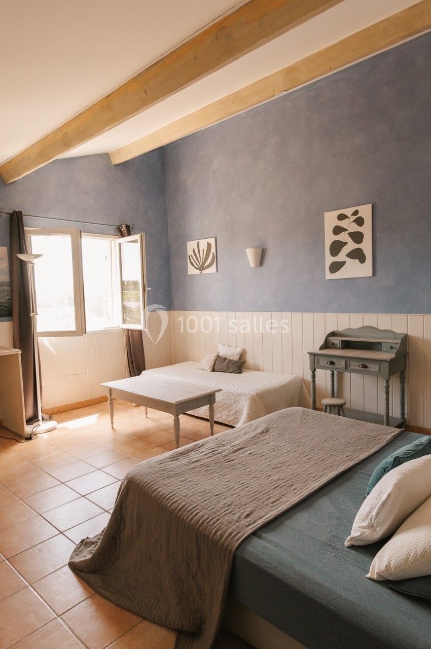 Chambre lumineuse avec deux lits simples, murs bleus, bureau, table basse et fenêtre ouverte sur l'extérieur.