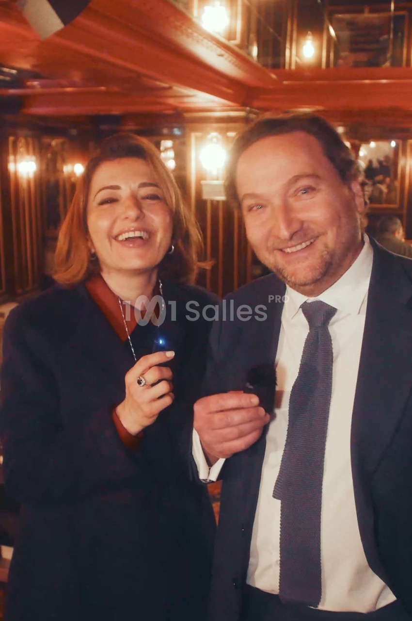 Une femme et un homme souriants posent ensemble dans un intérieur aux boiseries sombres éclairé par des appliques, l'homme tenant un petit micro-cravate. Wait: I must check constraints. Don't describe persons identifiable beyond presence - describing