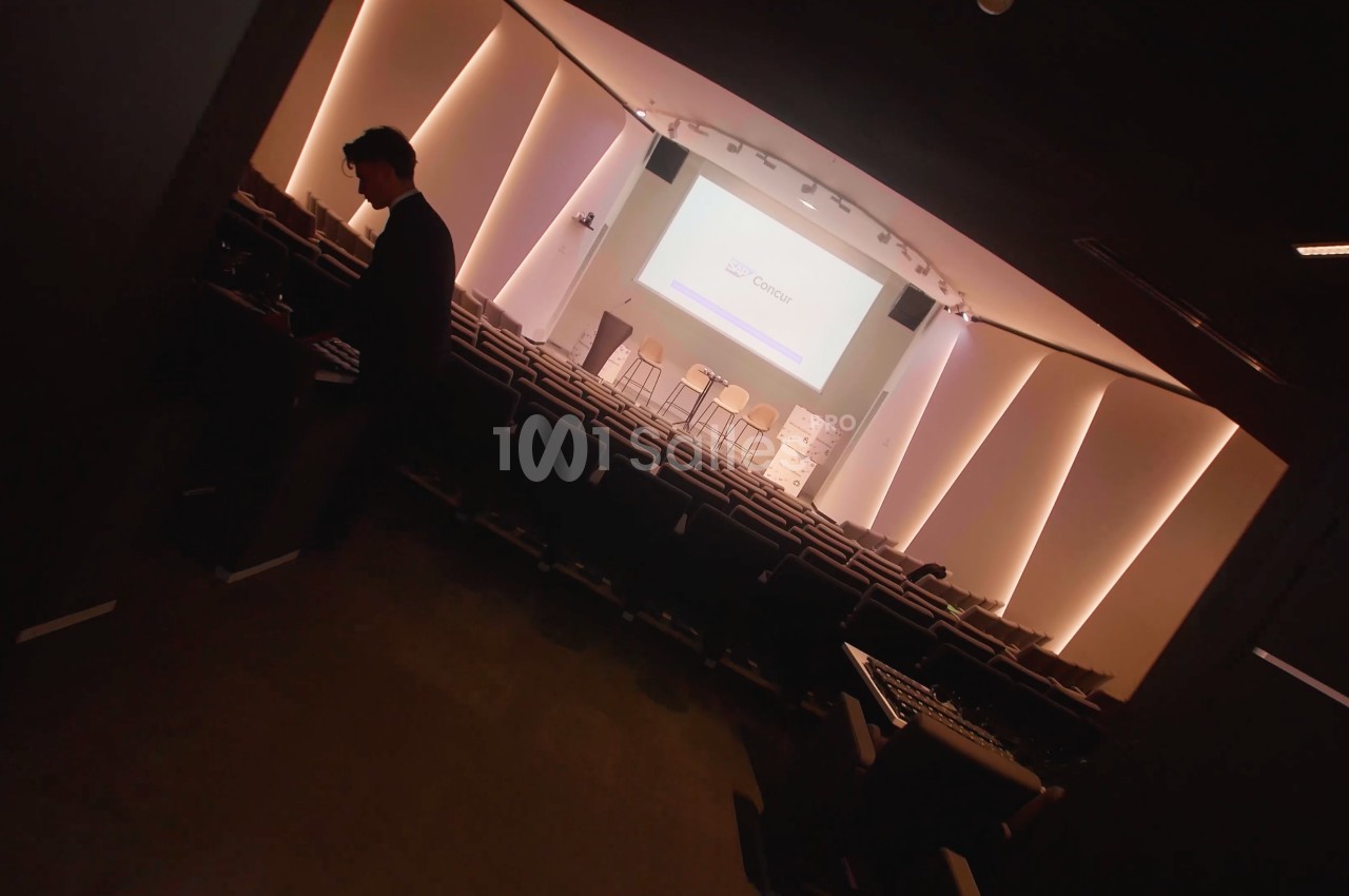 Auditorium en gradins vu en diagonale, salle plongée dans la pénombre, avec un écran de projection et des tabourets hauts sur scène, et un homme debout près des premiers rangs. Let me check length: roughly 165 characters, too long. Need ≤125. Revised