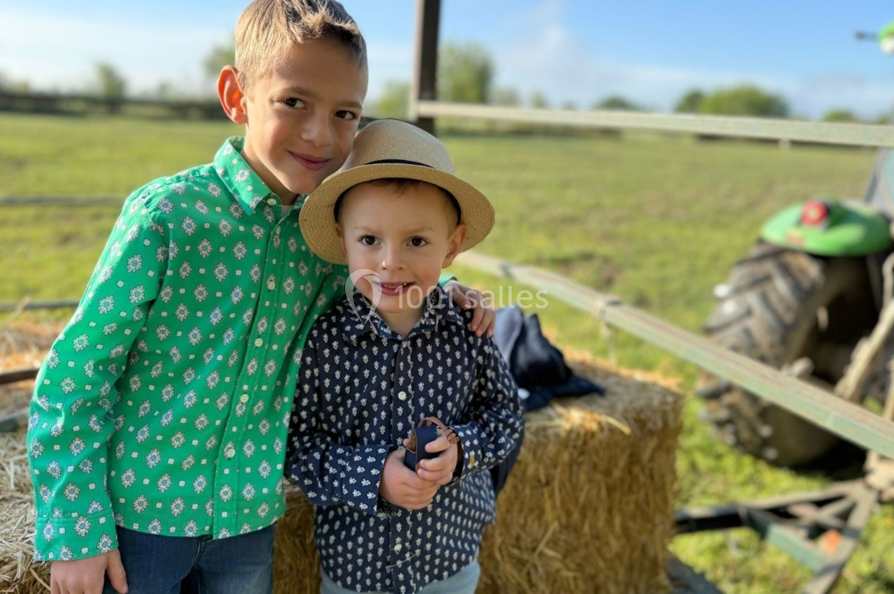 Deux enfants souriants posent près de bottes de foin dans un champ, avec un tracteur en arrière-plan.