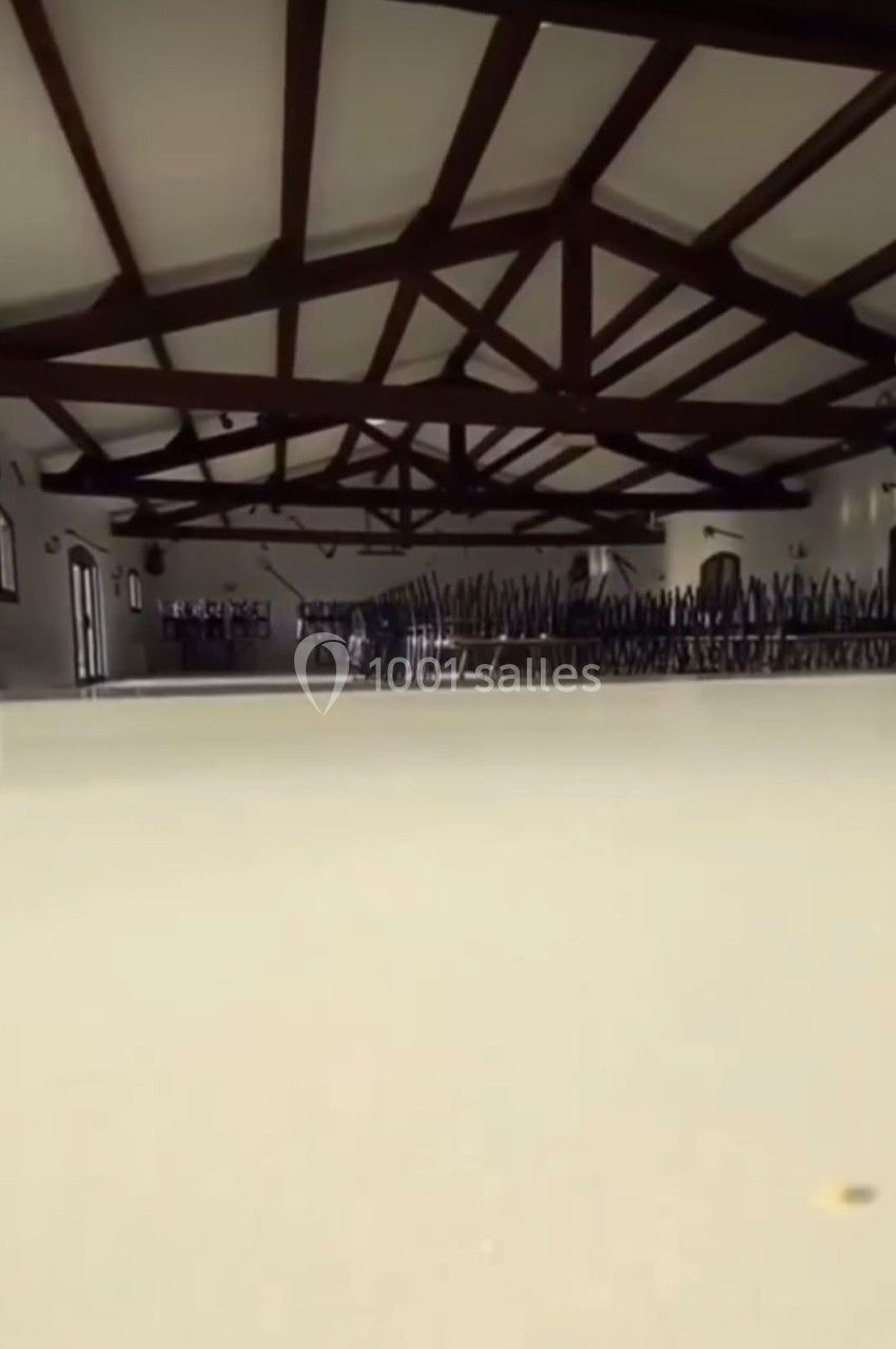 Salle vide avec plafond en poutres apparentes, chaises empilées dans le fond et lumière naturelle entrant par les fenêtres.
