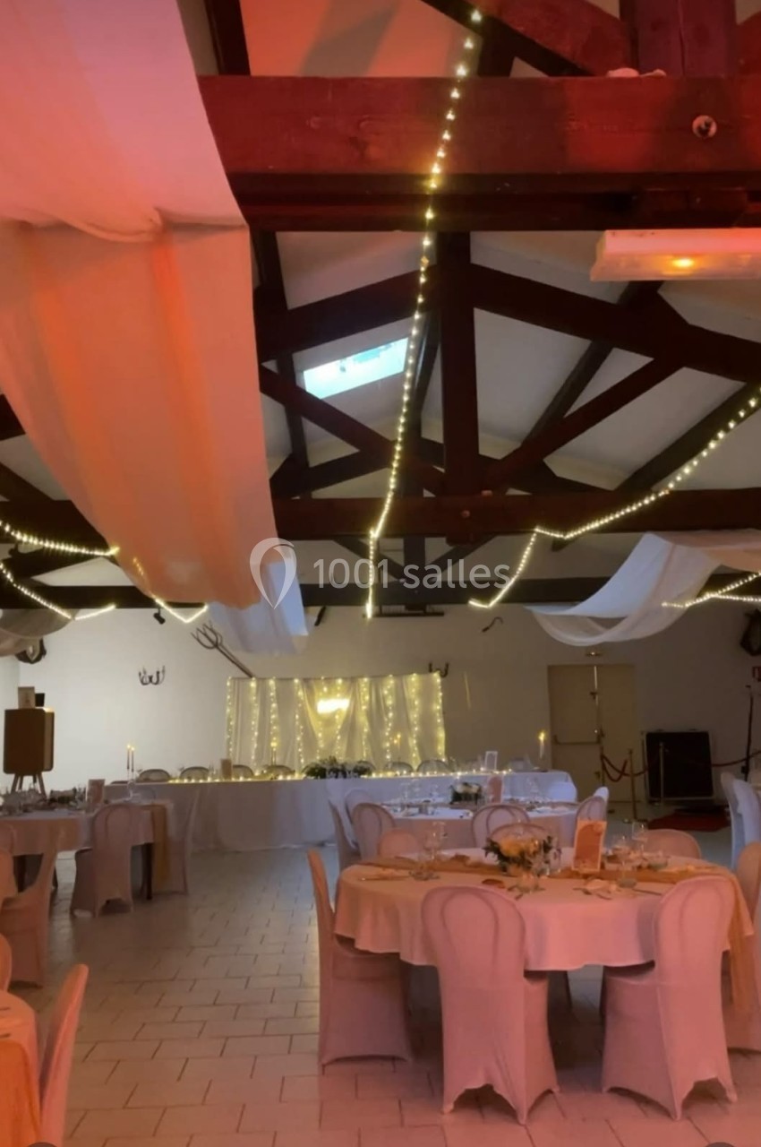 Salle de réception décorée avec des guirlandes lumineuses, des nappes blanches et des tables dressées pour un événement.
