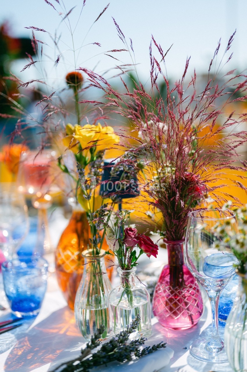 Vases colorés contenant des fleurs variées sur une table extérieure ensoleillée, décorée de verres et de lavande.