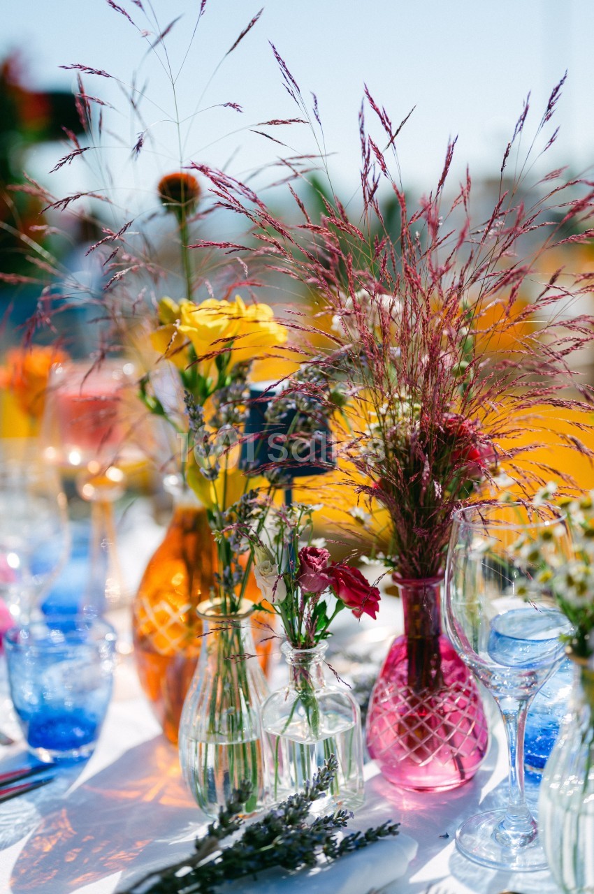 Vases colorés contenant des fleurs variées sur une table extérieure ensoleillée, décorée de verres et de lavande.