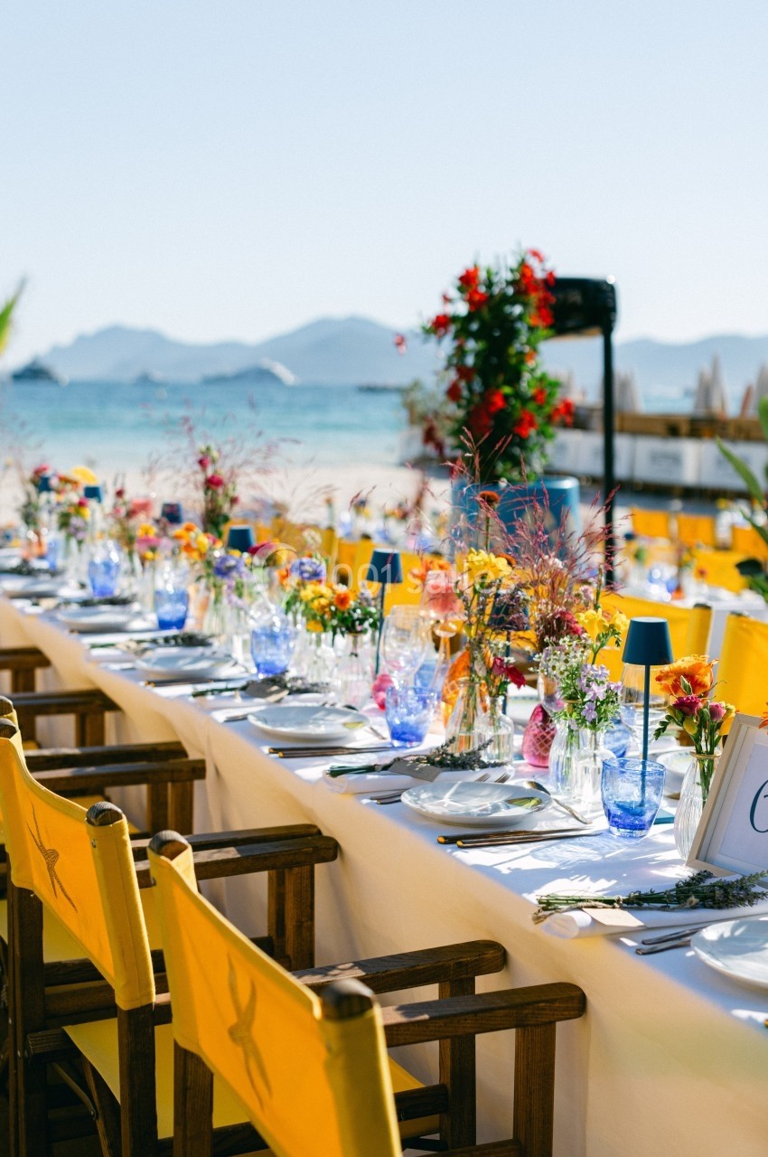 Table dressée en extérieur avec nappes blanches, chaises jaunes et décorations florales colorées, vue sur la plage et la mer.