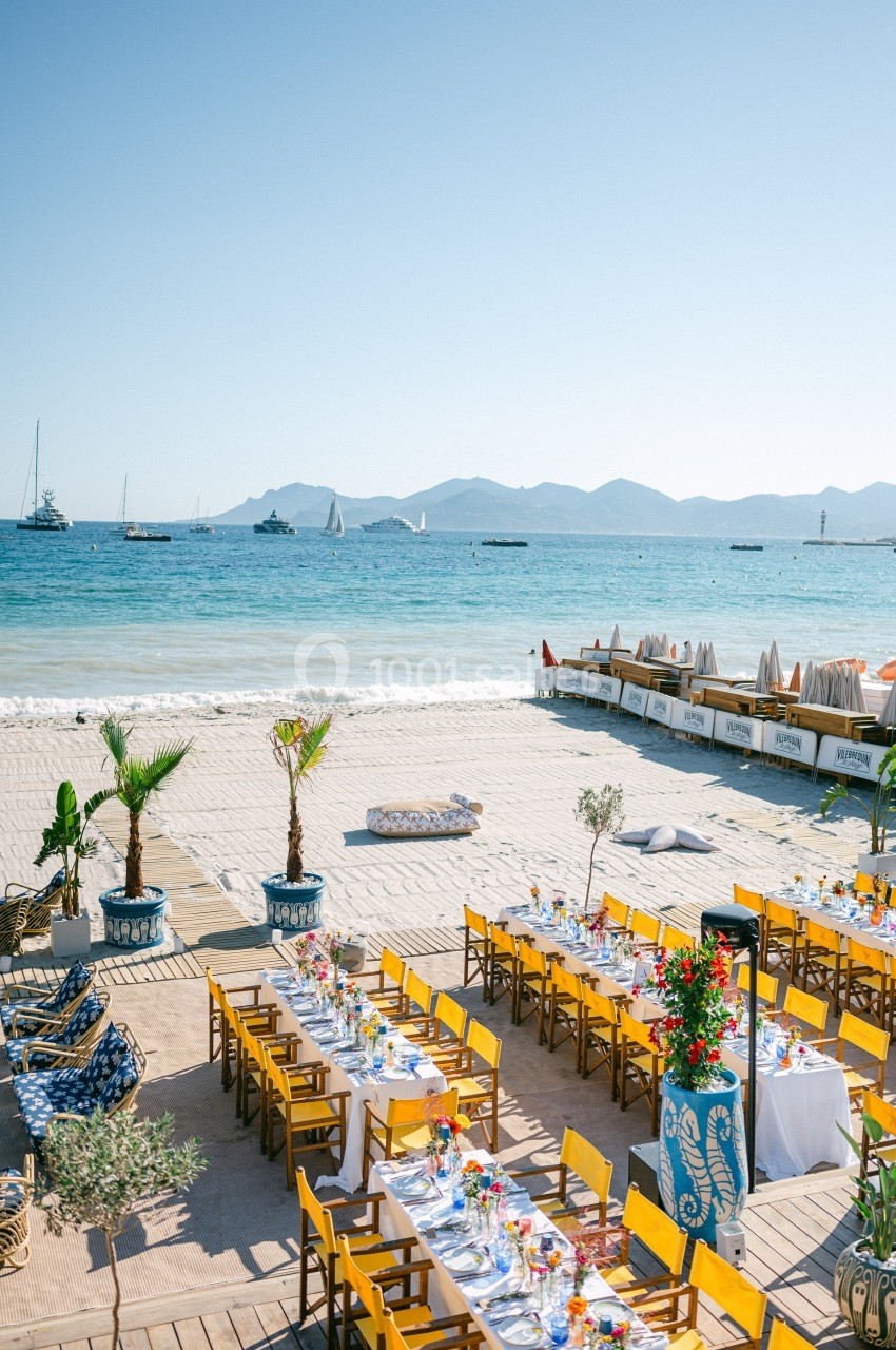 Tables dressées en extérieur face à la mer, avec des chaises jaunes, des palmiers en pot et des voiliers à l'horizon.