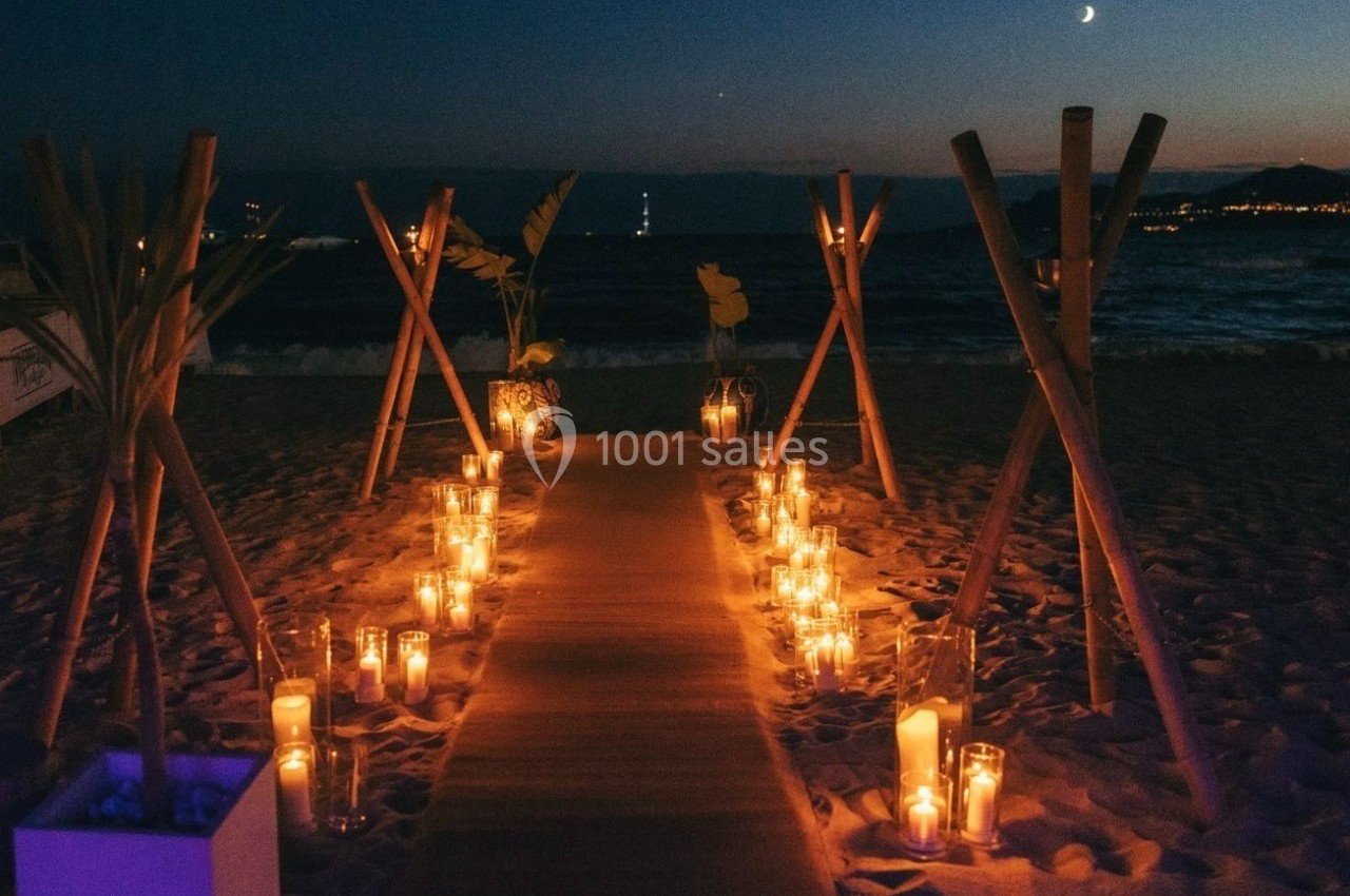 Allée éclairée par des bougies sur une plage au crépuscule, bordée de bambous et décorations tropicales.