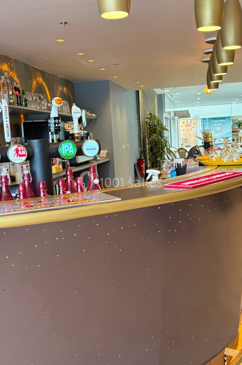 Comptoir de bar moderne avec tireuses à bière, bouteilles et luminaires suspendus, dans un espace lumineux.
