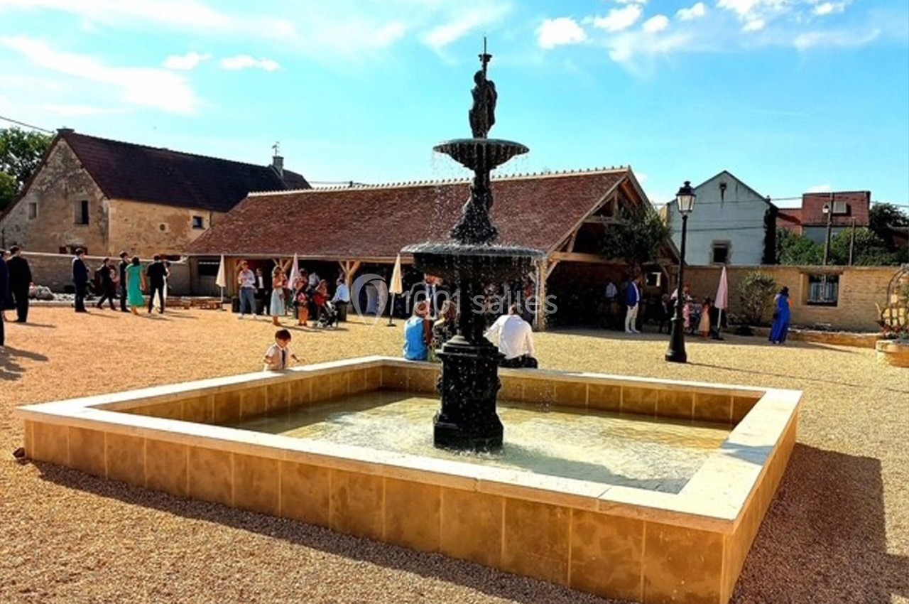 Fontaine centrale en pierre dans une cour ensoleillée, entourée de bâtiments traditionnels et de visiteurs.