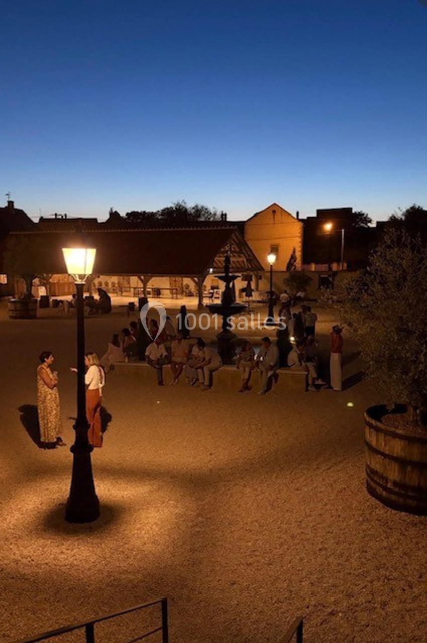 Place éclairée par des lampadaires au crépuscule, avec des personnes assises et debout près d'une structure en bois.