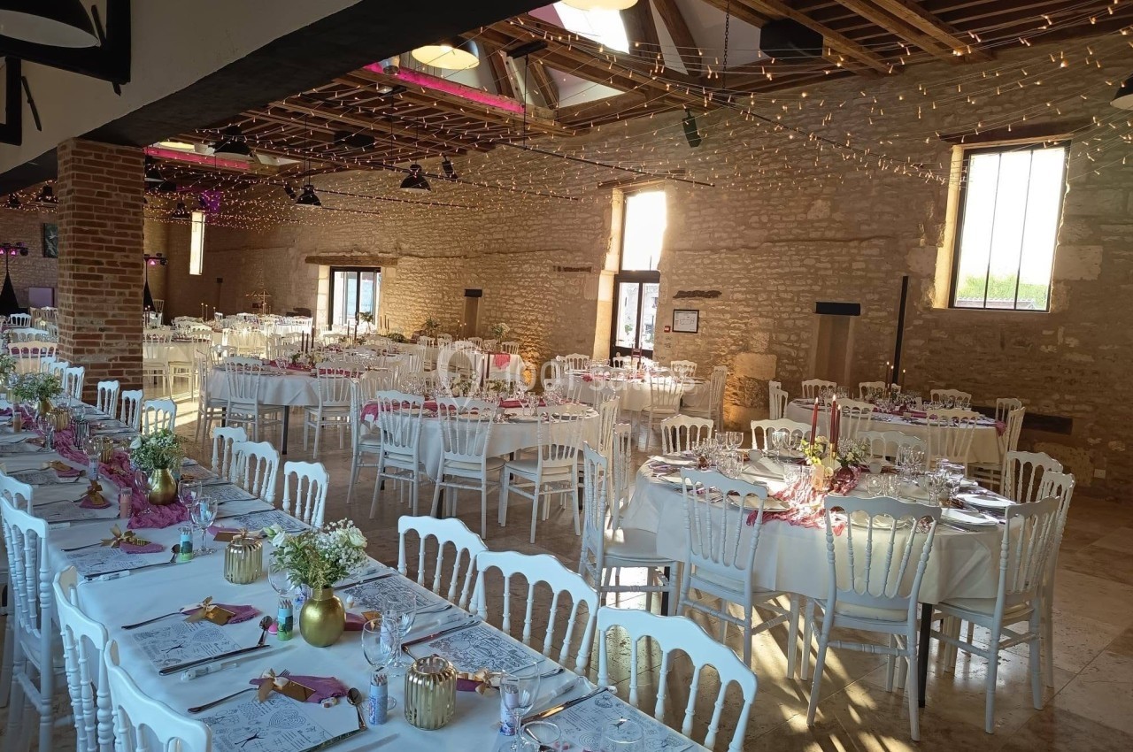Salle de réception décorée pour un événement, avec tables dressées, chaises blanches et guirlandes lumineuses suspendues.
