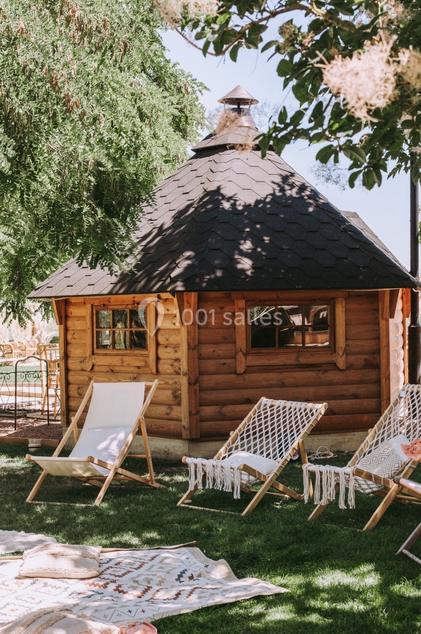Cabane en bois avec toit en bardeaux entourée de chaises longues et d'arbres dans un jardin ensoleillé.