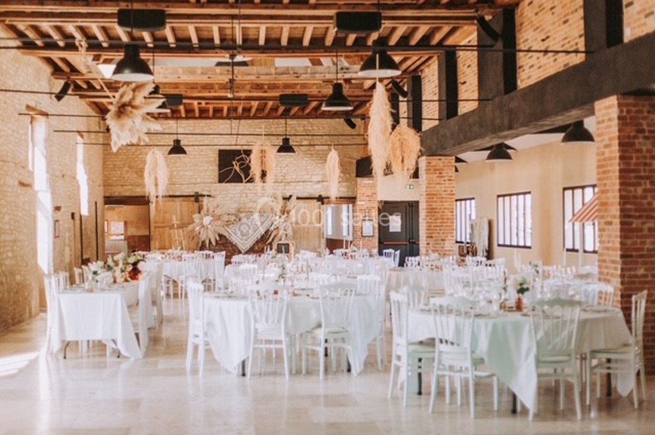 Salle de réception lumineuse avec tables dressées, chaises blanches et décorations suspendues en pampas.