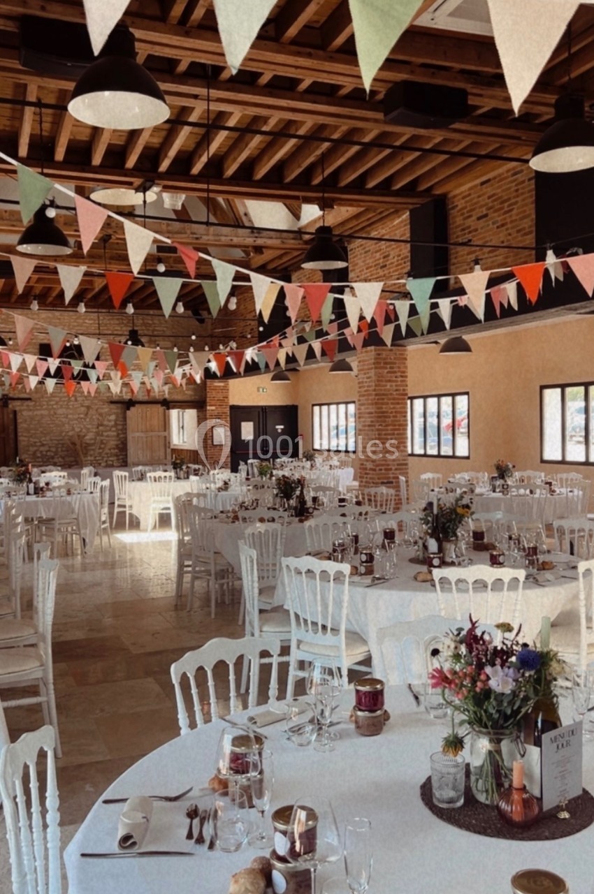 Salle de réception décorée avec des guirlandes colorées, des tables rondes dressées et des chaises blanches.