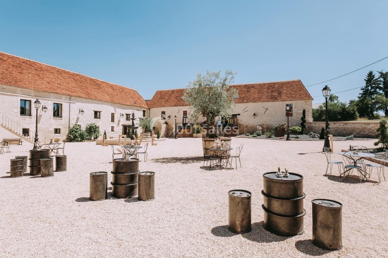 Cour extérieure d'une bâtisse en pierre avec tables, chaises et tonneaux métalliques sous un ciel dégagé.
