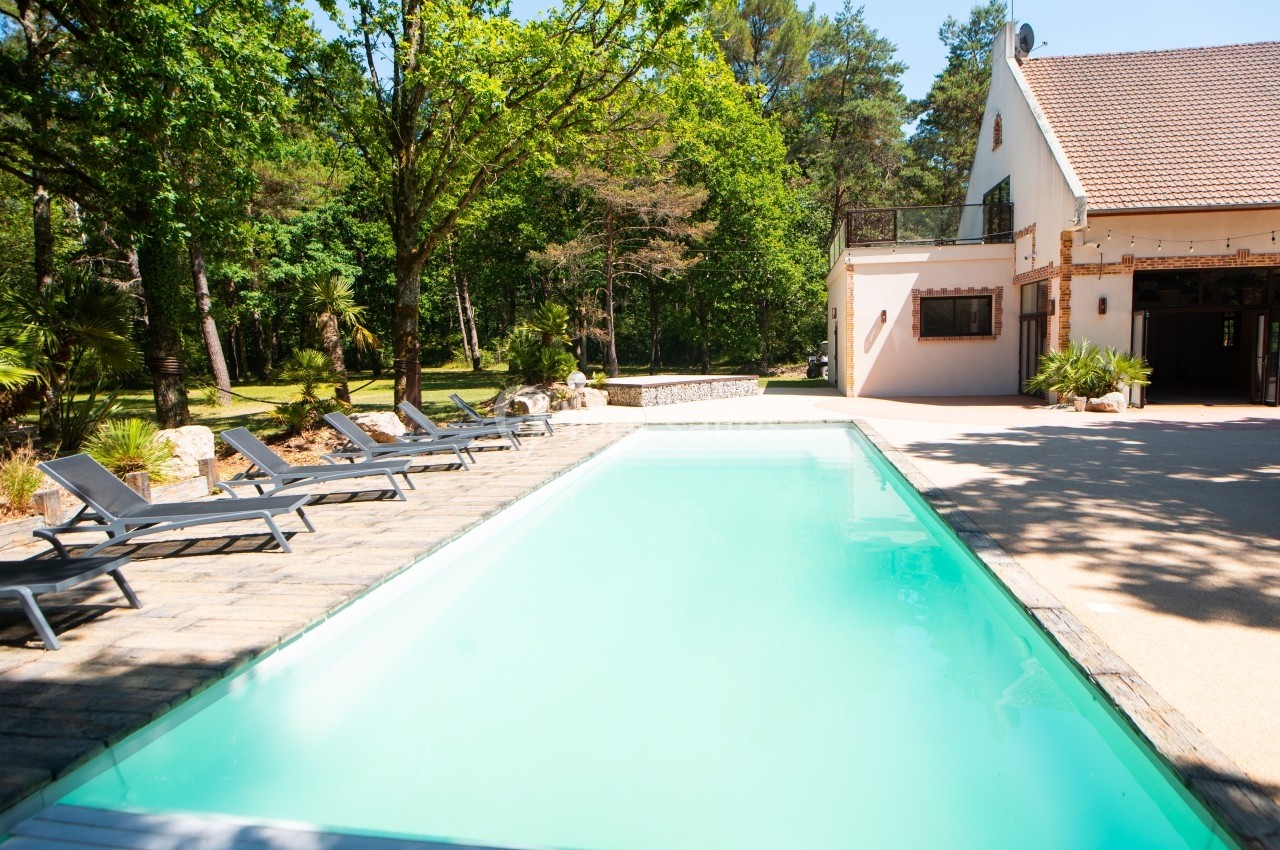 Piscine extérieure entourée de chaises longues, située dans un jardin arboré près d'une maison.