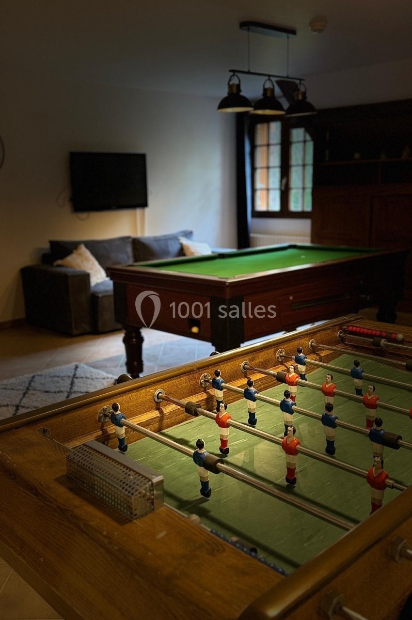 Baby-foot en premier plan avec des figurines colorées, billard et canapé en arrière-plan dans une pièce éclairée.