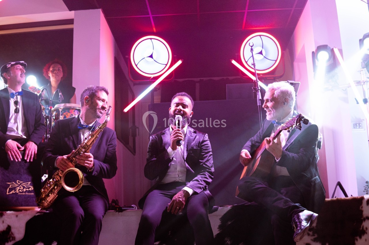 Un chanteur accompagné de musiciens jouant du saxophone, de la guitare et des percussions sur une scène éclairée.