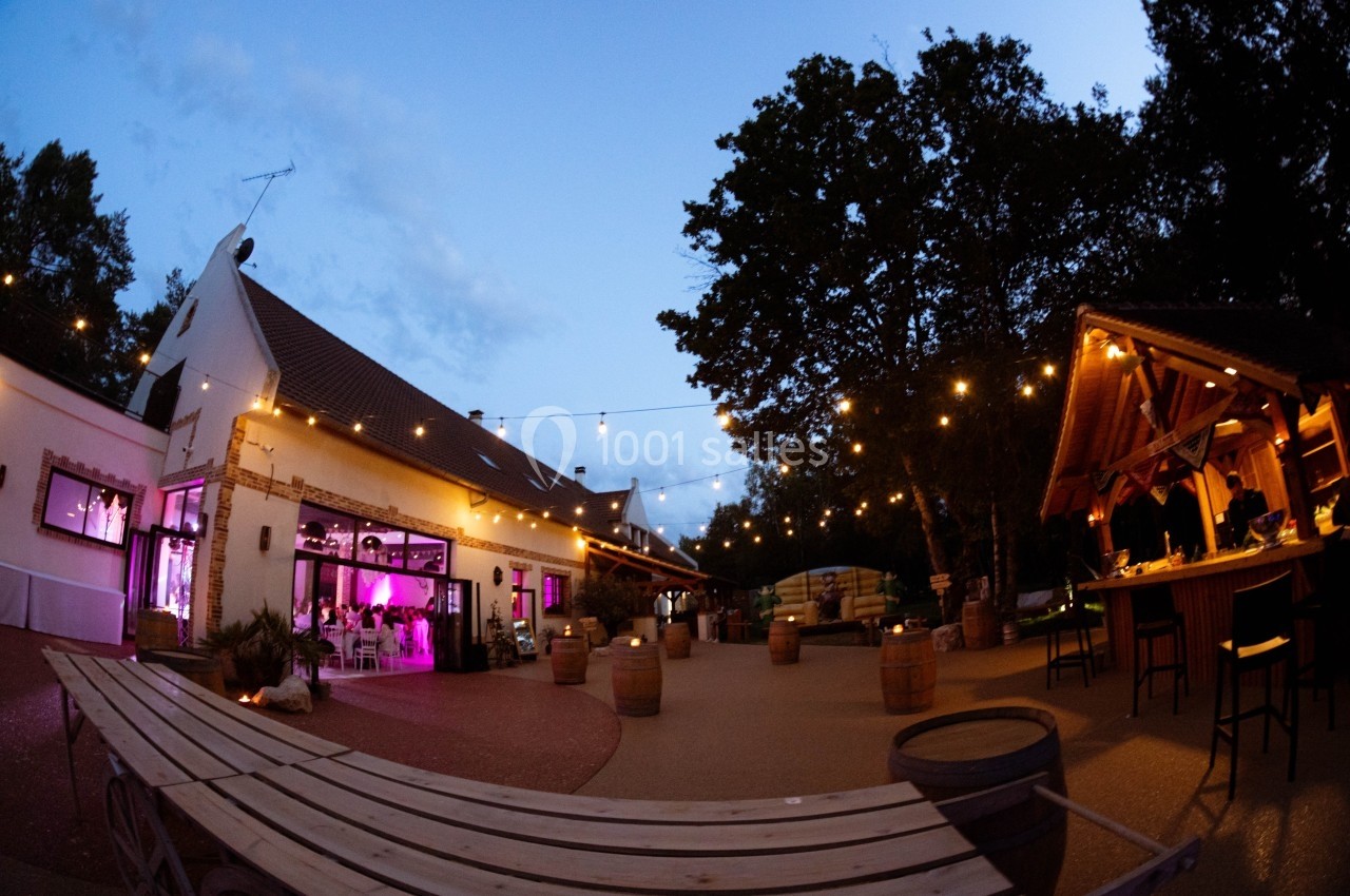 Terrasse éclairée en soirée avec guirlandes lumineuses, tables en bois et bâtiments au style champêtre.