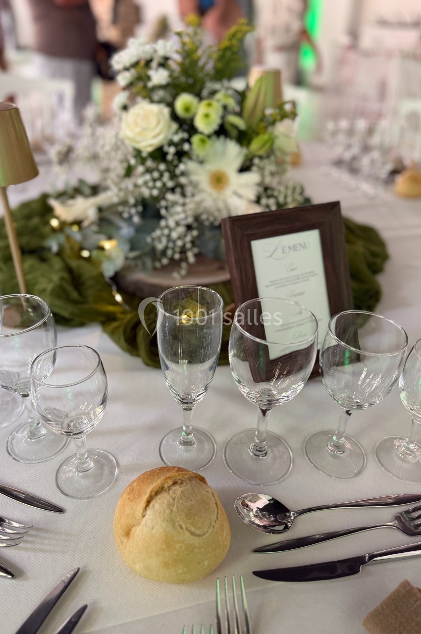 Table dressée avec vaisselle élégante, bouquet floral central, menu encadré et pain individuel sur une nappe blanche.