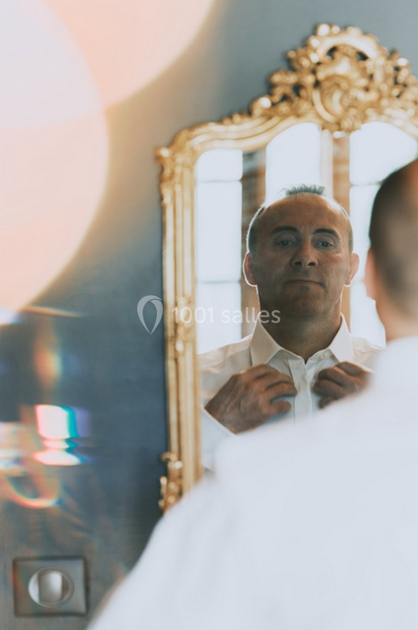 Un homme ajuste son col de chemise blanche devant un miroir doré dans une pièce lumineuse.