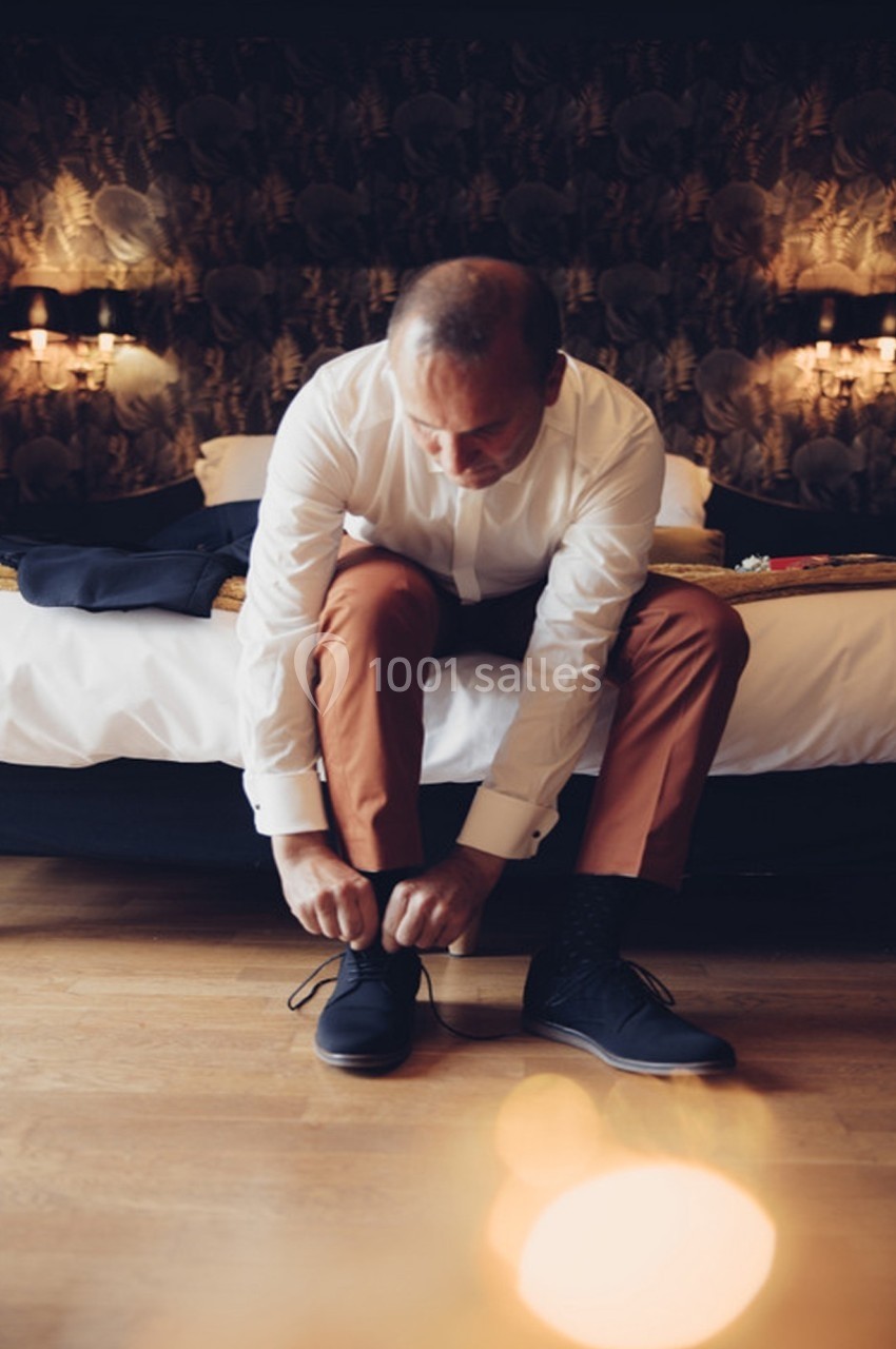 Un homme assis sur un lit noue ses chaussures noires dans une chambre à la lumière tamisée.