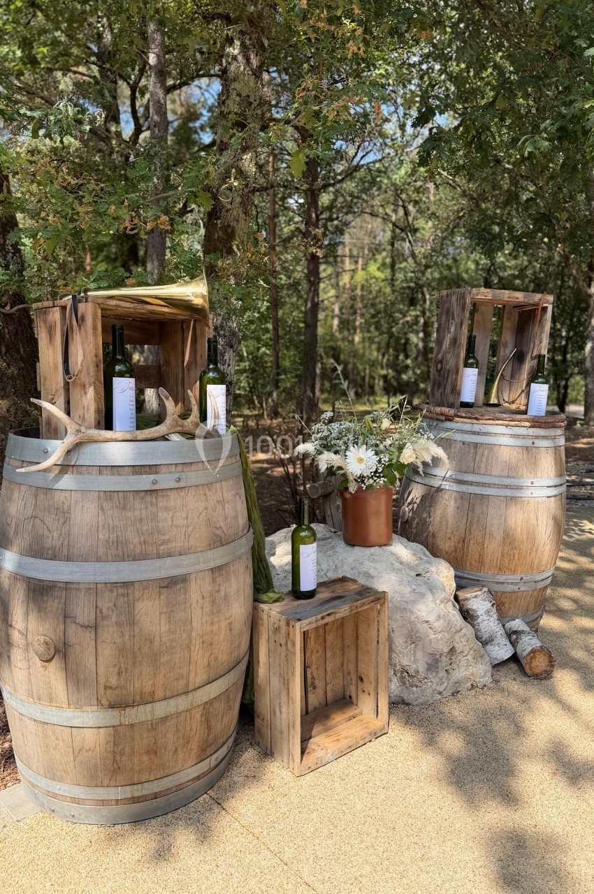 Deux tonneaux en bois décorés avec des bouteilles de vin, des fleurs et des caisses, dans un cadre extérieur boisé.
