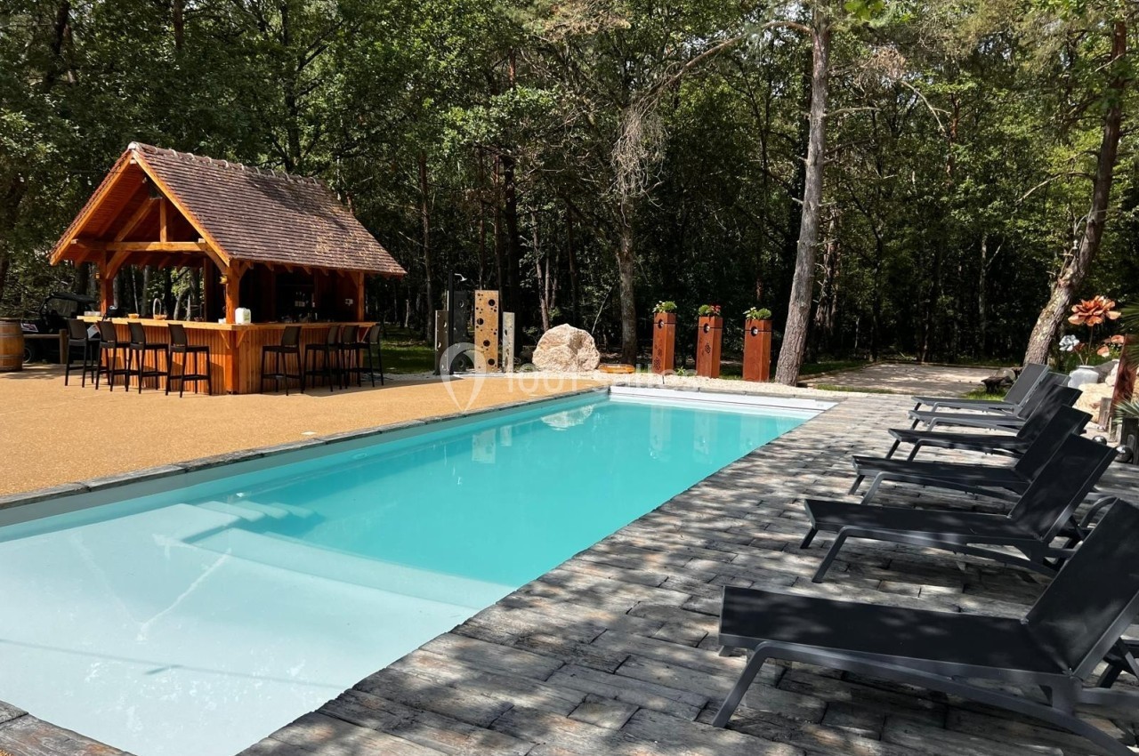 Piscine extérieure entourée de chaises longues noires, avec un kiosque en bois et une forêt en arrière-plan.