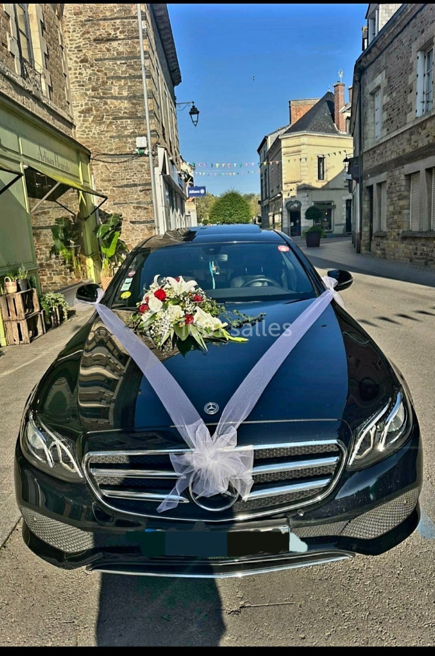 Voiture noire décorée de rubans blancs et d'un bouquet de fleurs, stationnée dans une rue pavée.