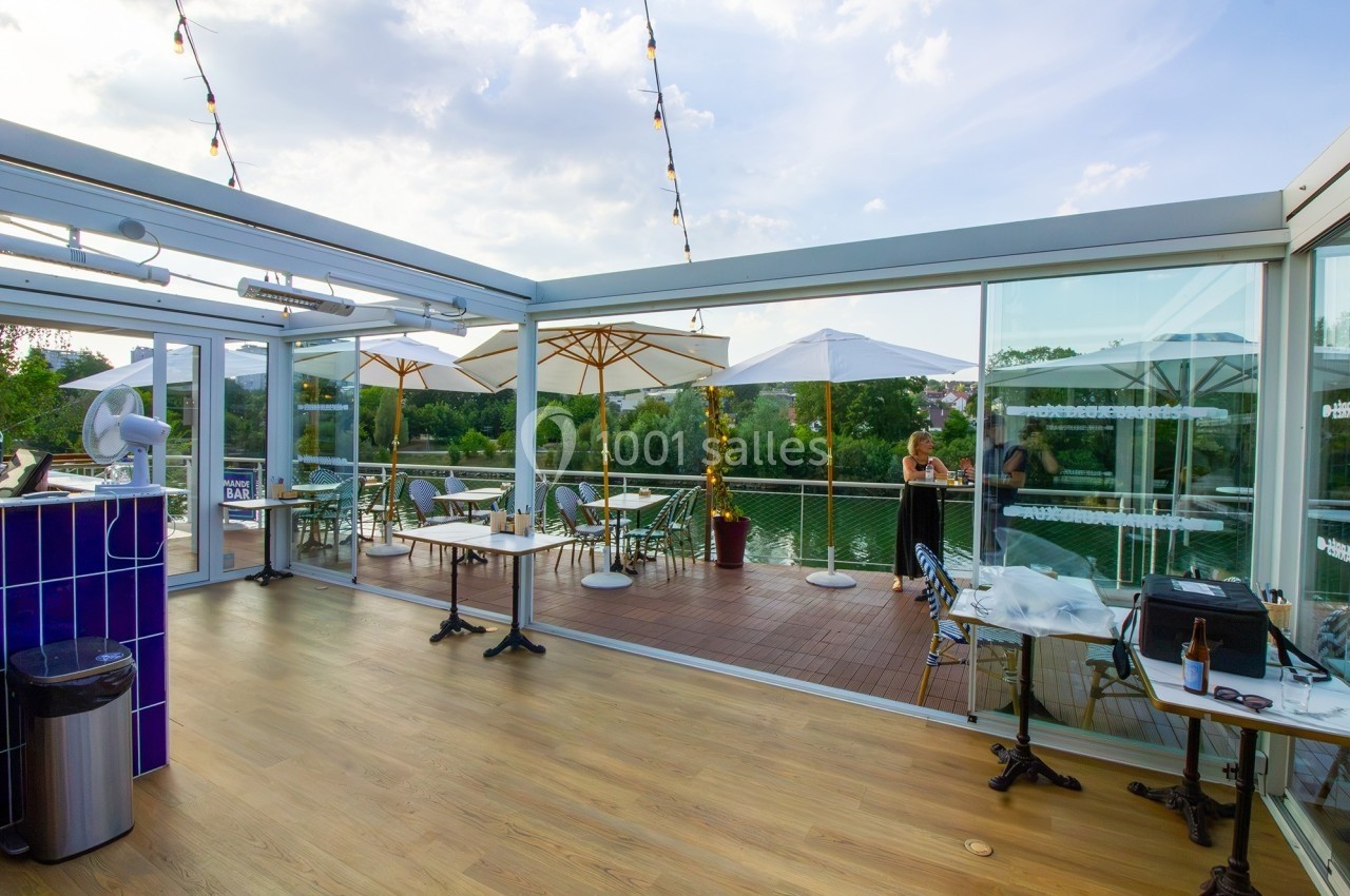 Terrasse extérieure avec tables, chaises, parasols et vue sur un plan d'eau, entourée de verdure.