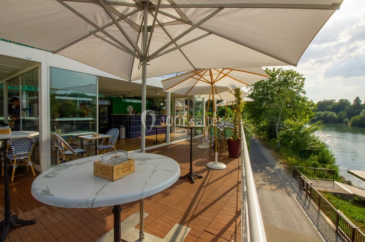 Terrasse ensoleillée avec tables et parasols, donnant sur une rivière et entourée de verdure.