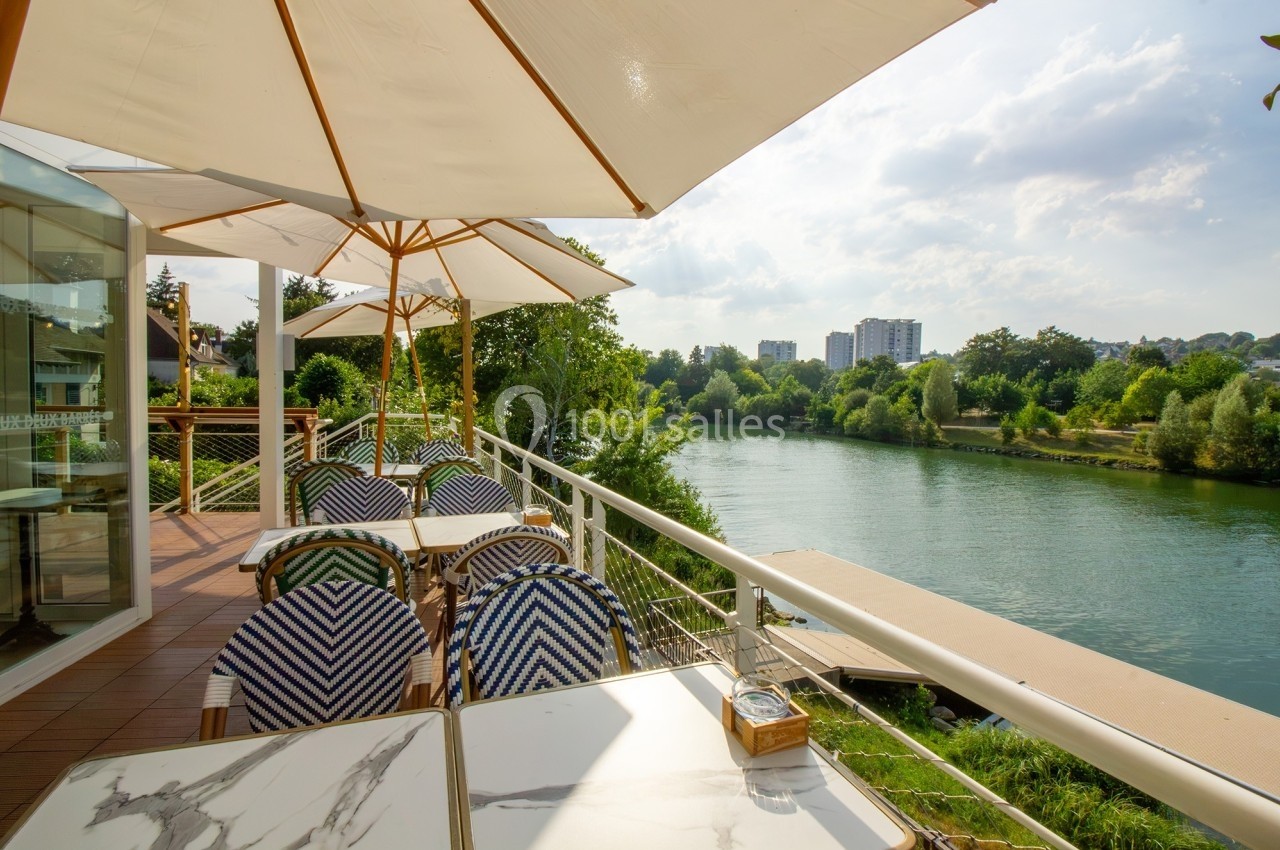 Terrasse d'un restaurant avec des tables et des chaises sous des parasols, offrant une vue sur une rivière et des arbres.