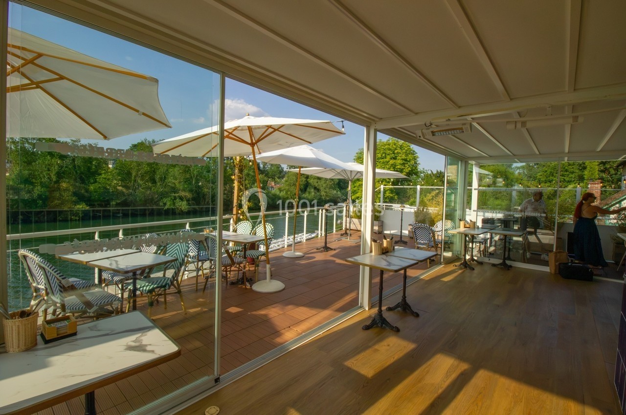 Terrasse ensoleillée d'un restaurant au bord de l'eau, avec parasols blancs et mobilier extérieur élégant.