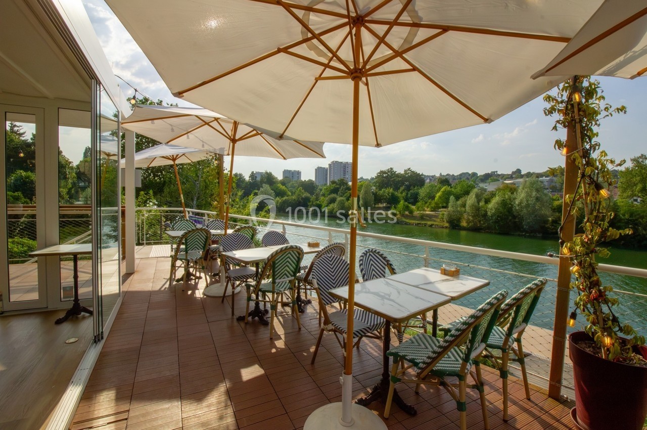 Terrasse ensoleillée avec tables, chaises et parasols, donnant sur une rivière bordée de verdure.