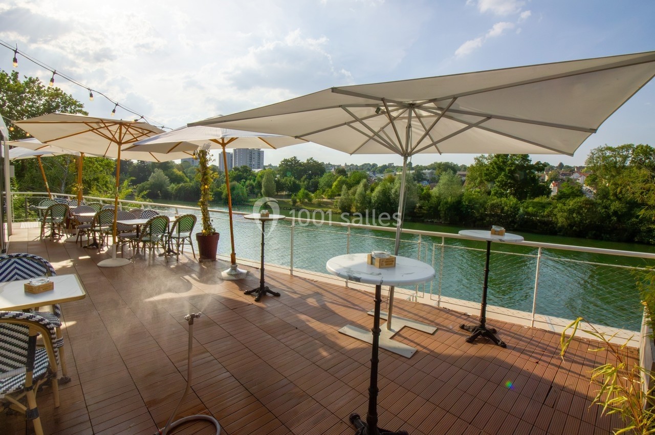 Terrasse en bois avec tables, chaises et parasols, offrant une vue sur une rivière bordée de végétation.