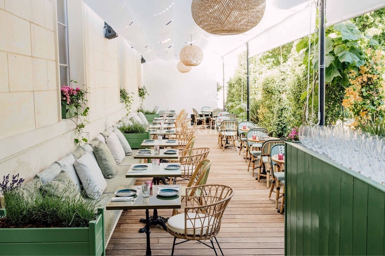 Terrasse couverte dressée avec tables alignées, chaises en rotin, couverts bleus et murs végétalisés.